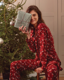 Žena sedi pored jelke sa poklonom u ruci obučena u novogodišnju pidžamu